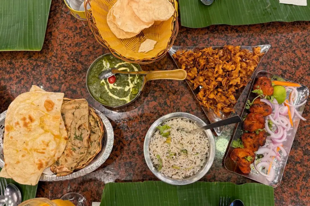 A vibrant spread of Indian dishes on a dark granite table, featuring various items such as naan bread, a bowl of saag paneer, crispy fried pakora, a serving of rice, and chicken tandoori garnished with onions and lime.
