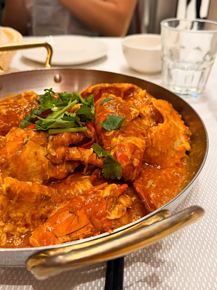 A metallic bowl filled with chili crab, garnished with fresh coriander, is placed on a table. In the background, a glass of water and a bowl can be partially seen.