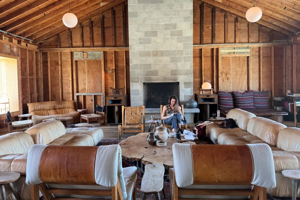 A cozy and rustic interior of a lodge with wooden beams, a stone fireplace, and eclectic seating arrangements. A woman sits comfortably in the room, engaged in an activity.