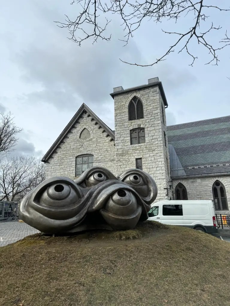 A contemporary sculpture featuring three large, eye-like shapes sits in front of a stone church building with gothic architectural elements under a cloudy sky.