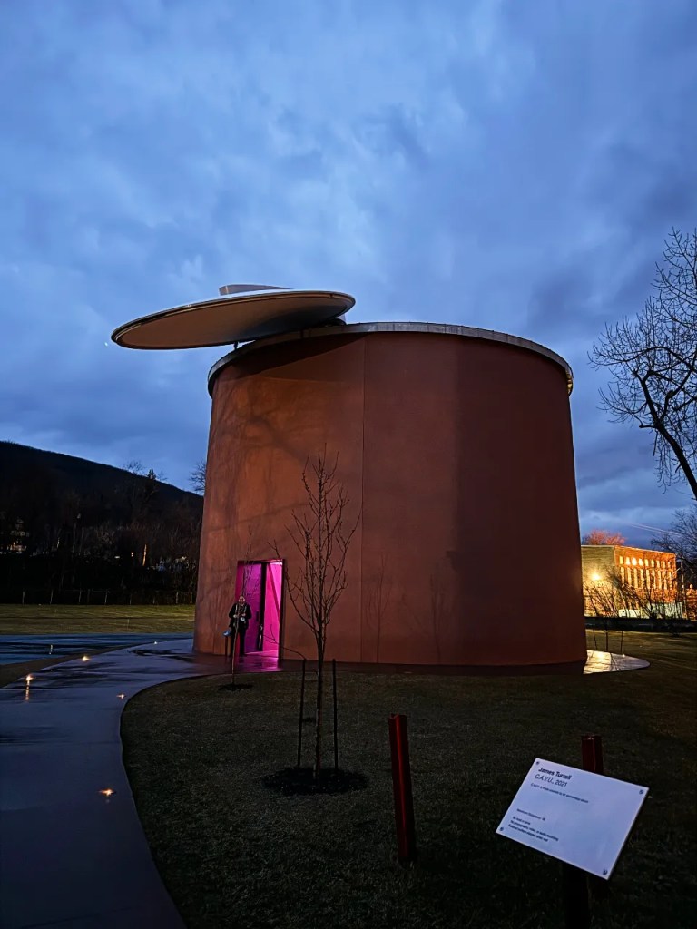 Night view of James Turrell's CAVU installation, featuring a cylindrical structure with a large disc on top, illuminated doorway, and landscaped surroundings.