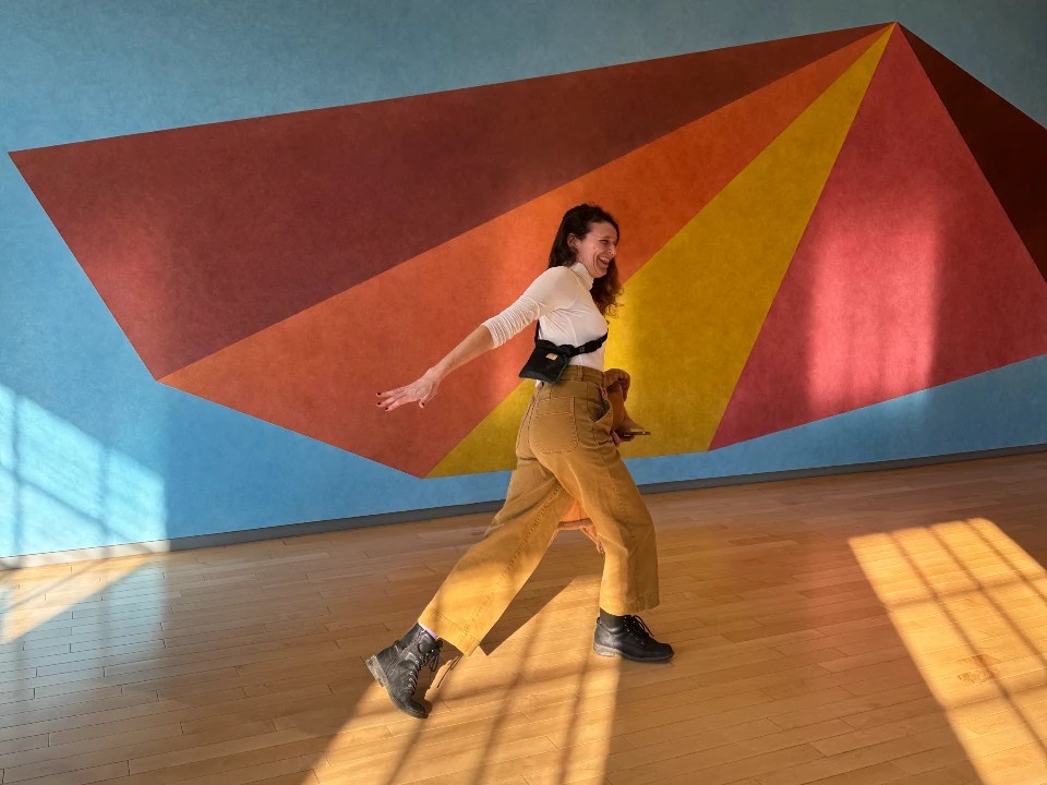 A person joyfully posing in front of a colorful geometric mural in a brightly lit indoor space.