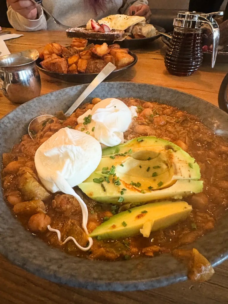 A close-up of a hearty breakfast dish featuring poached eggs and avocado, served in a gray bowl alongside potatoes and syrup in the background.