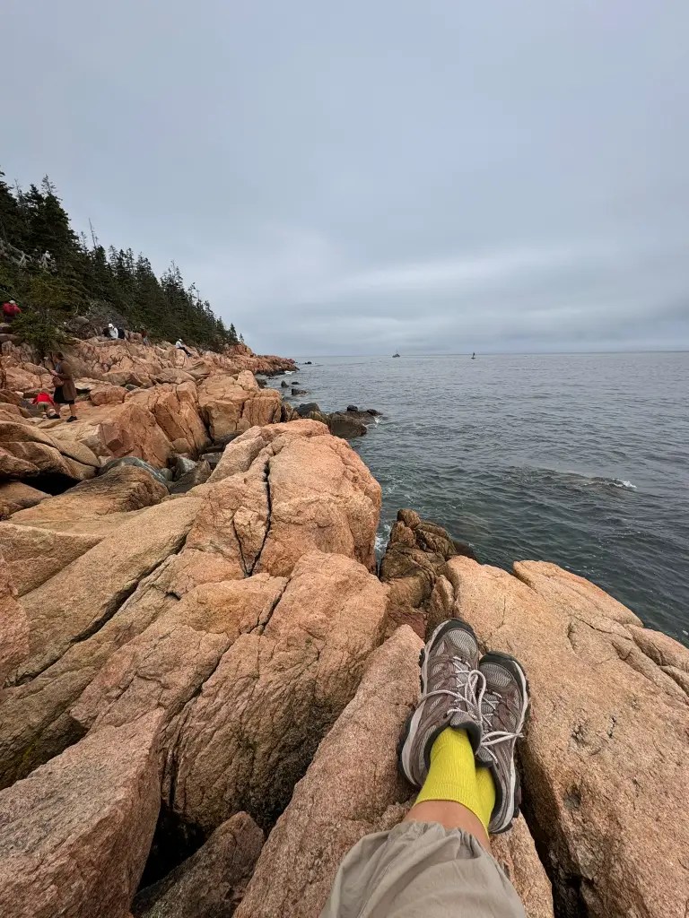 A view from above of a hiker's feet resting on rocky shorelines beside calm water, with overcast skies and trees in the background.