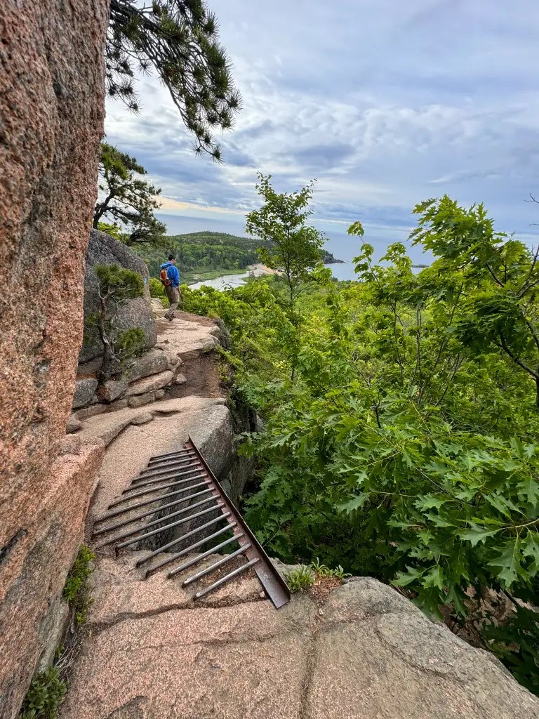 A hiker navigating a rocky trail with safety railings, surrounded by lush green foliage and distant views of the coast under a partly cloudy sky.