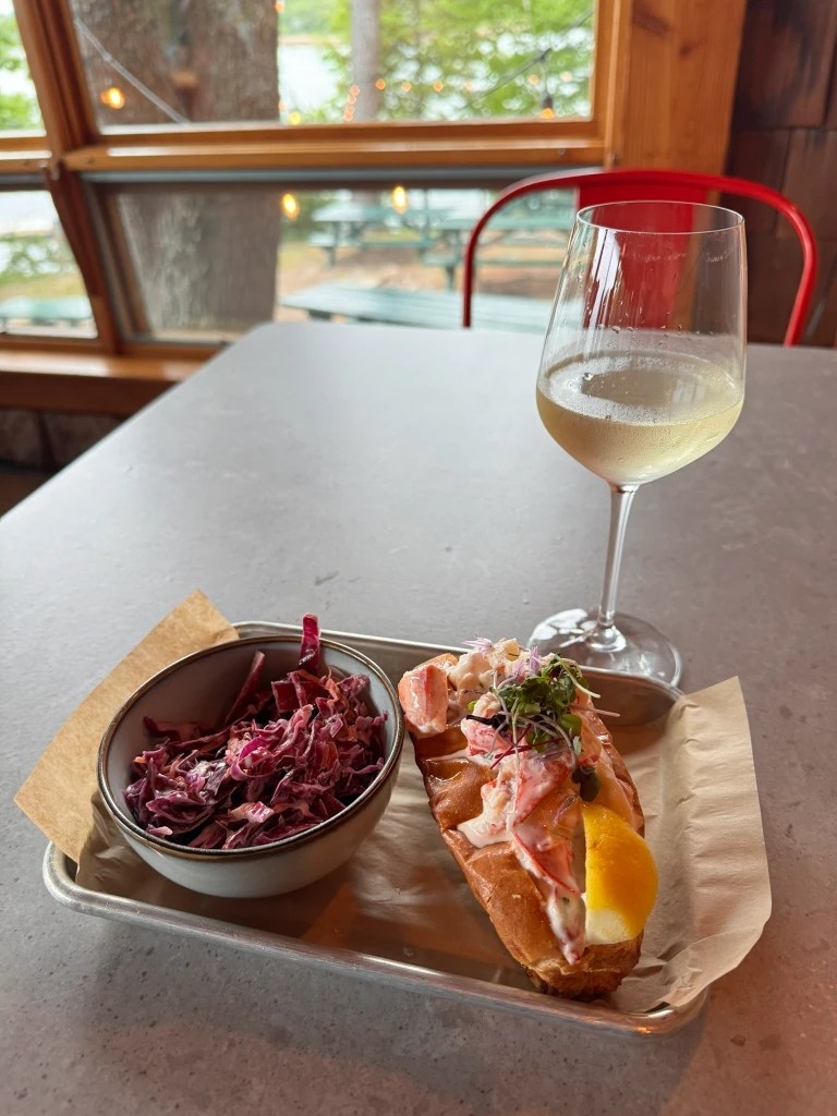 A lobster roll served on a toasted bun with a lemon wedge, accompanied by a bowl of red cabbage slaw and a glass of white wine, set against a backdrop of wooden interior and greenery outside.