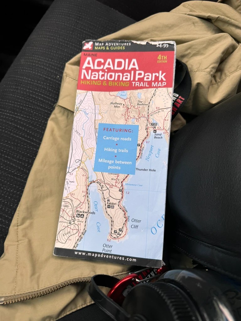 A close-up of an Acadia National Park hiking and biking trail map, featuring details about carriage roads and hiking trails, resting on a seat partially covered by a jacket.