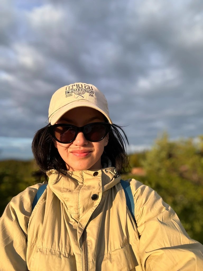 A person wearing sunglasses and a beige jacket with a cap stands outdoors, smiling against a backdrop of partly cloudy skies.