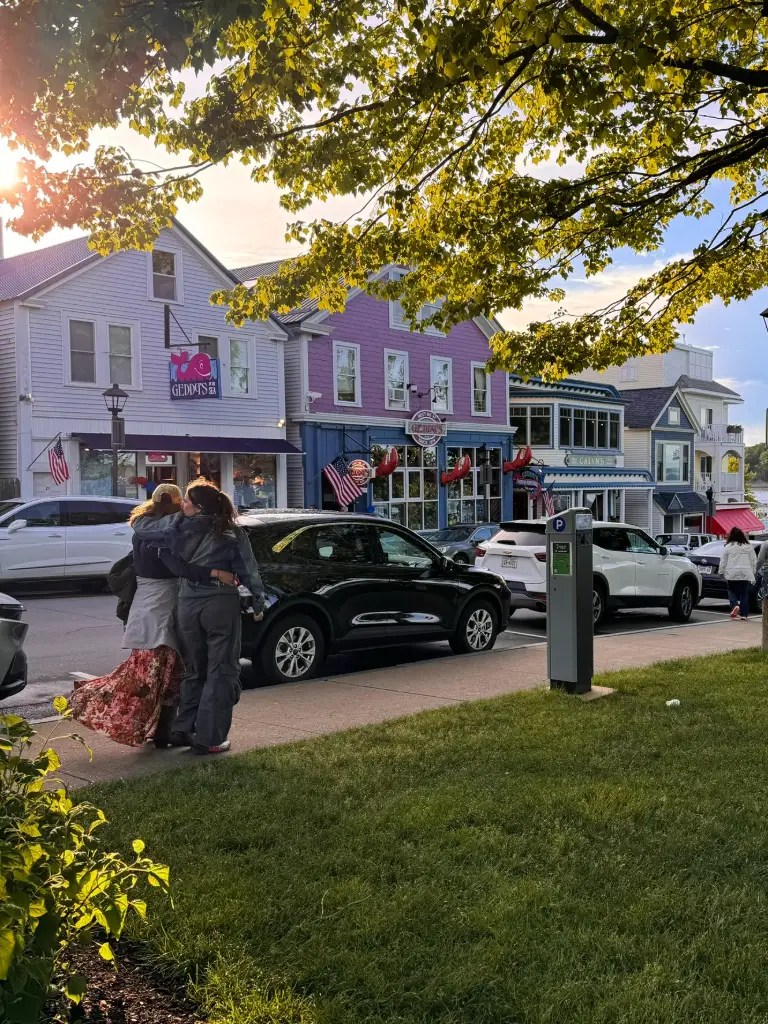 A pair of people standing close together on a sidewalk, enjoying their time in a scenic downtown area with colorful buildings and American flags. Cars are parked along the street, and the sun is setting in the background, casting a warm glow.