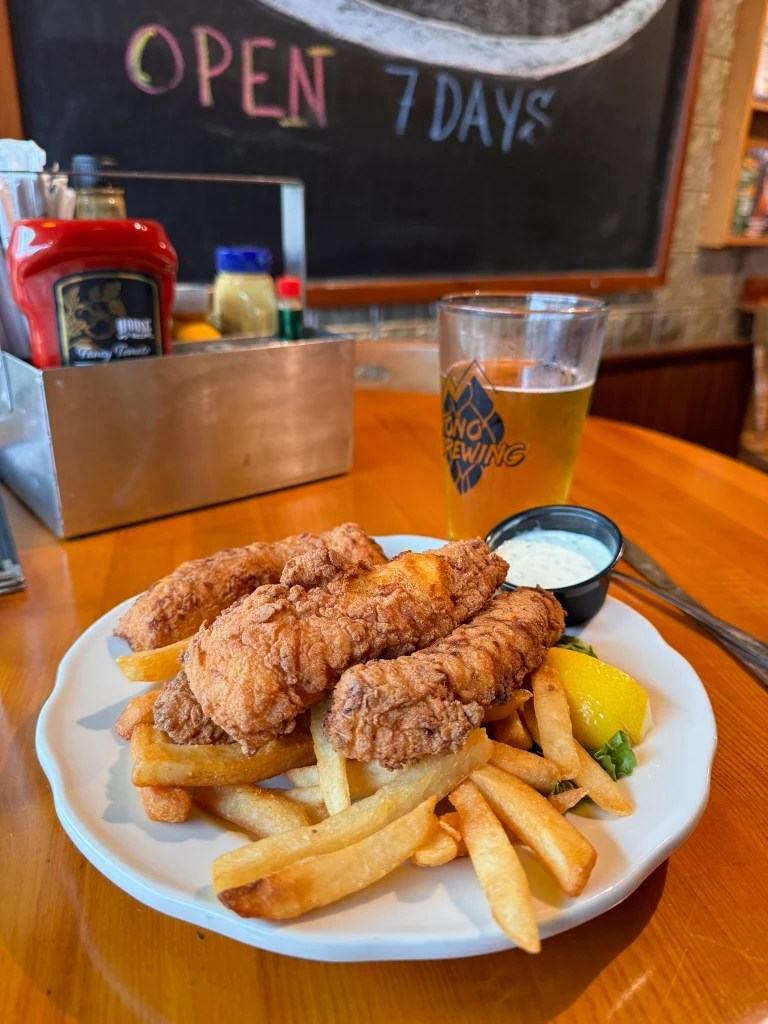 A plate of fried fish and chips with three crispy fish fillets, golden French fries, a slice of lemon, and a small cup of dipping sauce, set on a wooden table with a beer glass in the background.