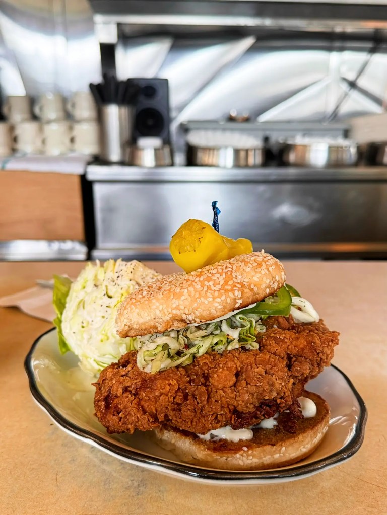A close-up of a crispy fried chicken sandwich featuring lettuce and pickles, nestled in a sesame seed bun, with a side of coleslaw and a metal counter in the background.