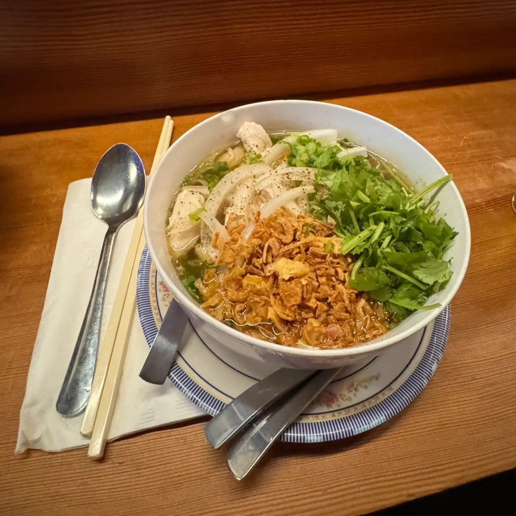 A bowl of Vietnamese chicken pho topped with herbs, fried onions, and sliced onions, served with chopsticks and a spoon on a wooden table.