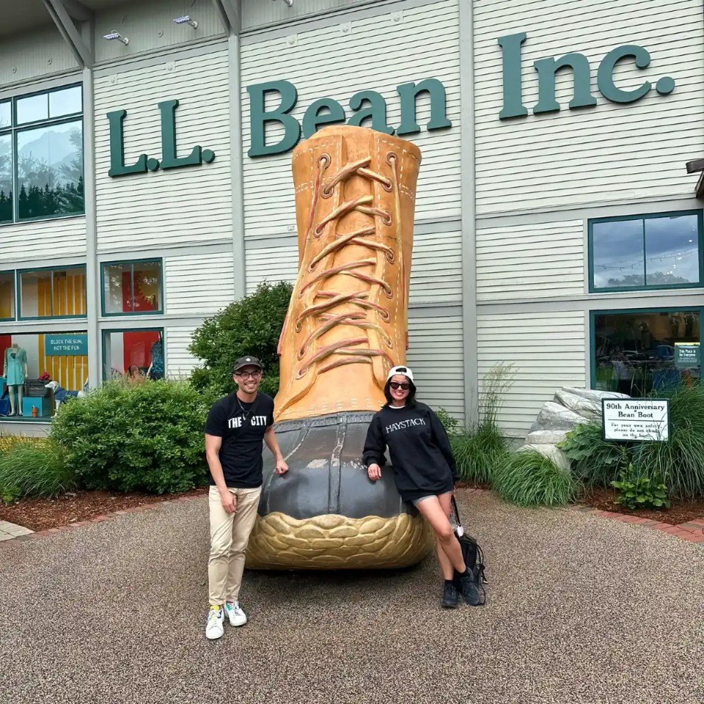 Two people standing next to a giant L.L. Bean boot in front of an L.L. Bean store, with greenery and a sign visible.