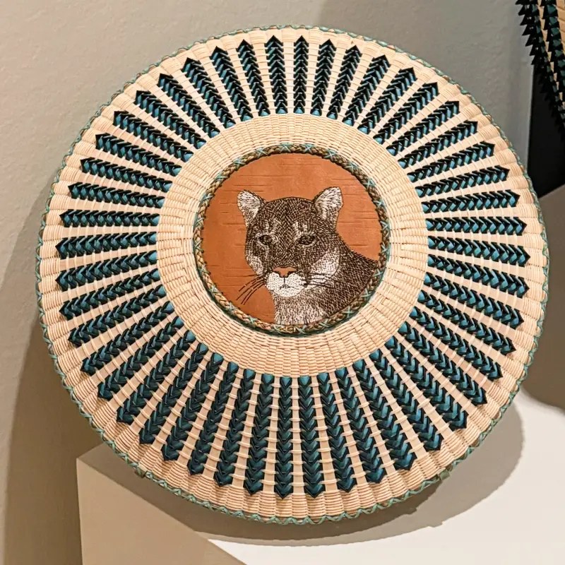A decorative woven basket featuring an embroidered face of a mountain lion in the center, surrounded by a radial pattern of blue and natural-colored fibers.