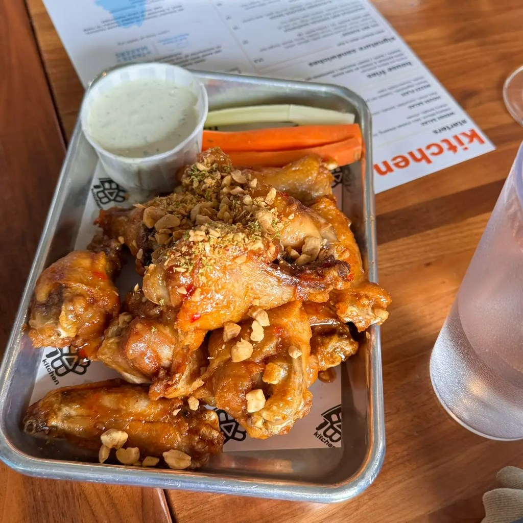 A close-up of a plate of crispy chicken wings garnished with crushed peanuts and spices. Accompanying the wings are sticks of celery and carrot, along with a small cup of ranch dressing. The setting appears to be a casual restaurant.