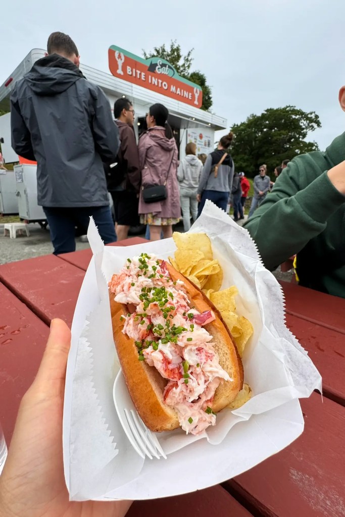A close-up of a lobster roll topped with chives, served with potato chips, held in hand with a food truck serving lobster rolls in the background.