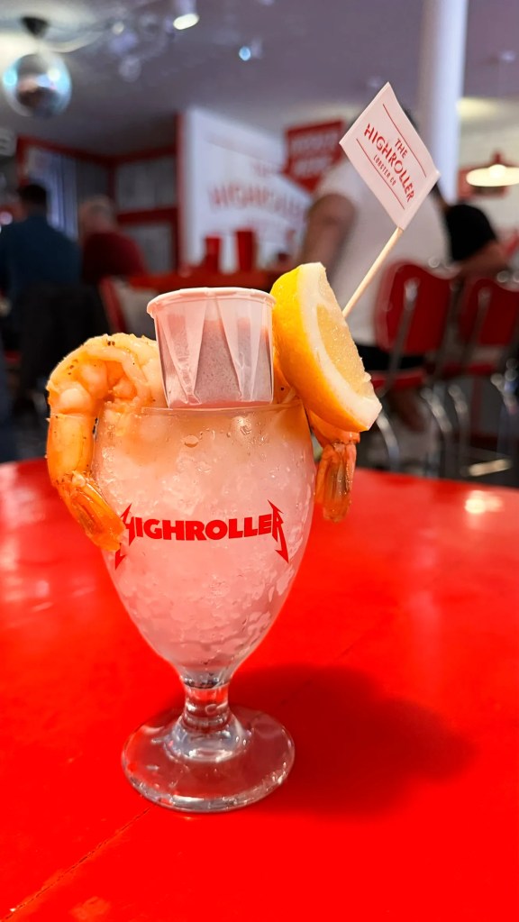A seafood cocktail garnished with shrimp, a lemon wedge, and a flag from The Highroller Lobster Co, served in a decorative glass on a bright red tabletop.