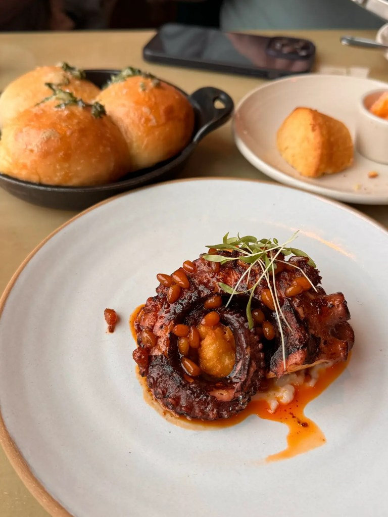 A plate featuring tender, grilled octopus topped with pine nuts and sauce, served alongside soft, golden rolls in a small dish.
