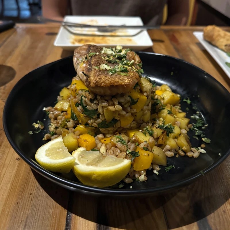 A dish featuring a fillet of seabass on top of a mound of grains and diced butternut squash, garnished with herbs and served with lemon wedges, on a wooden table.