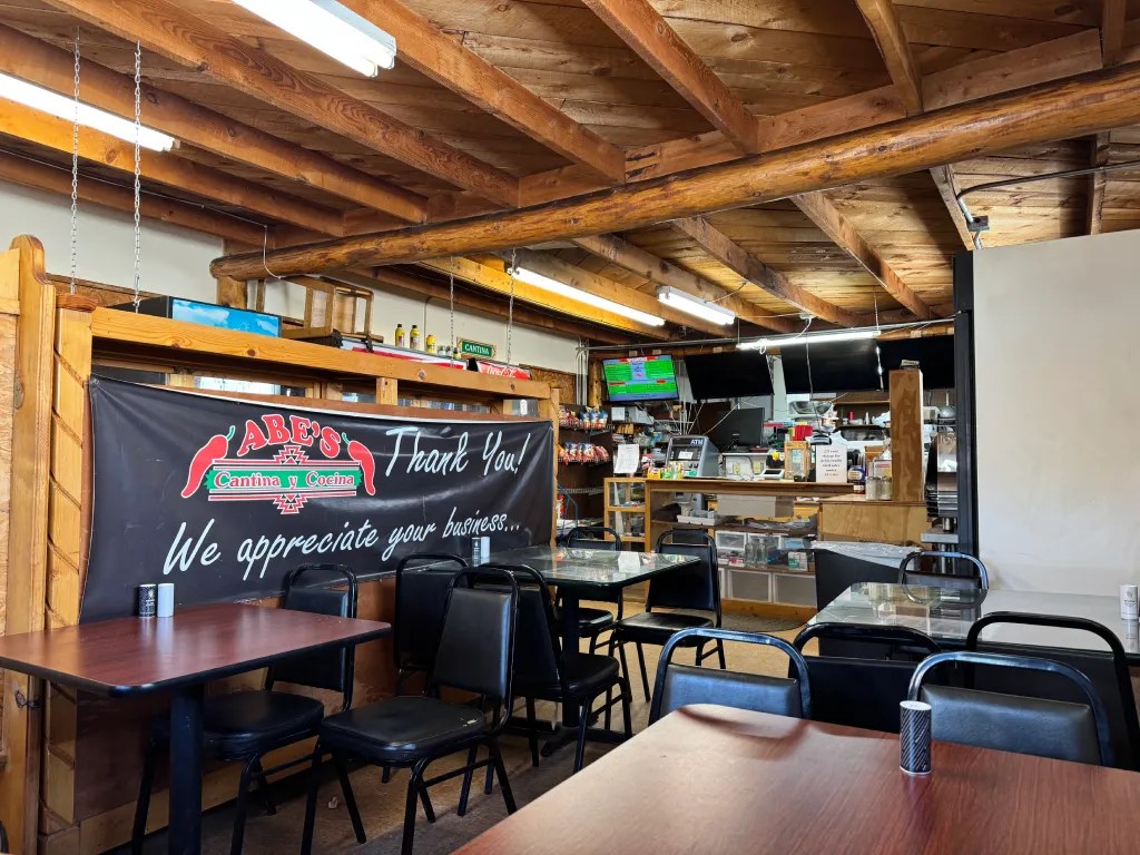 Interior of Abe's Cantina y Cocina, featuring wooden beams and tables. A banner thanking customers hangs prominently, and the service area with a cash register and menu items is visible in the background.