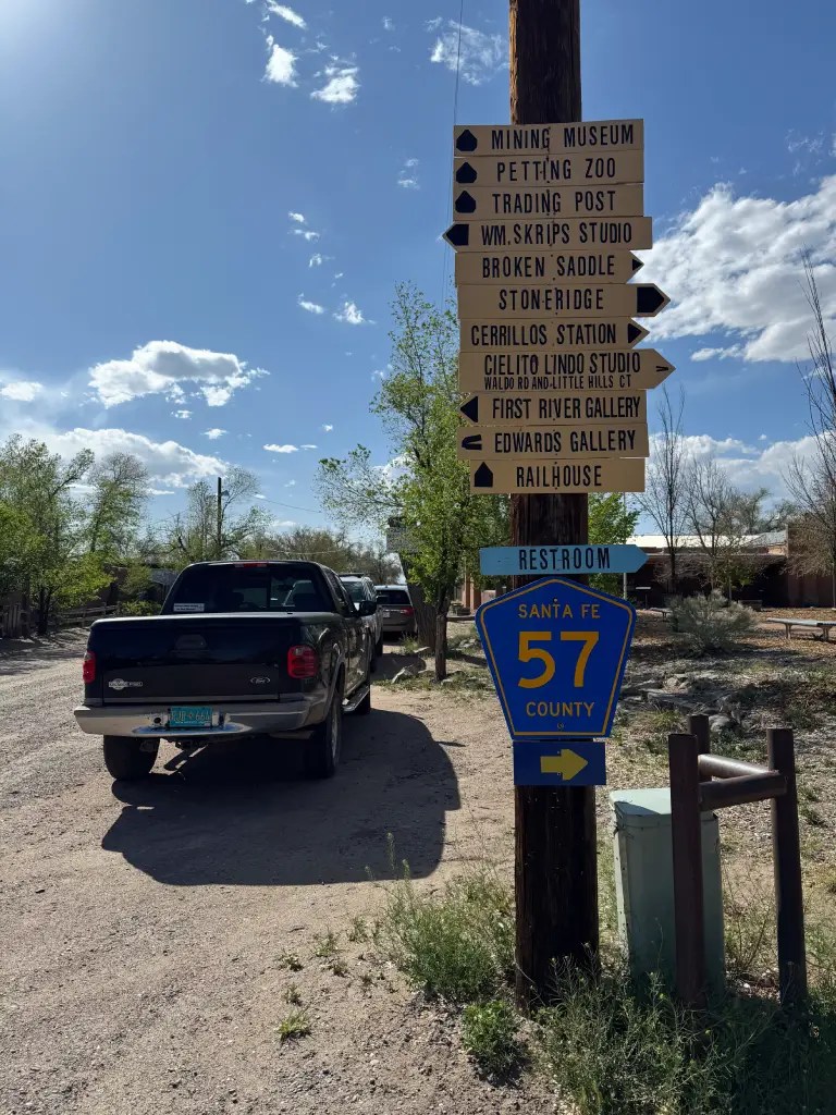 A wooden signpost displaying directional arrows and names of various attractions like 'Mining Museum,' 'Petting Zoo,' and 'Trading Post' alongside a blue county road sign indicating 'Santa Fe 57'. In the background, a pickup truck is parked on a dirt road under a partly cloudy sky.