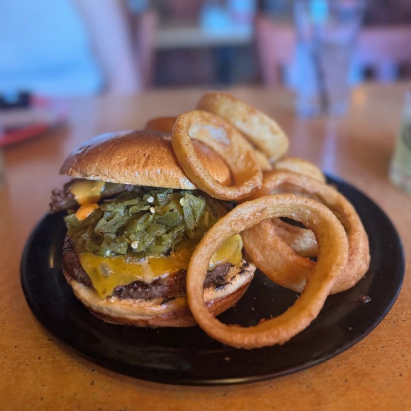A juicy burger topped with melted cheese and green chiles, accompanied by a side of onion rings, served on a black plate.