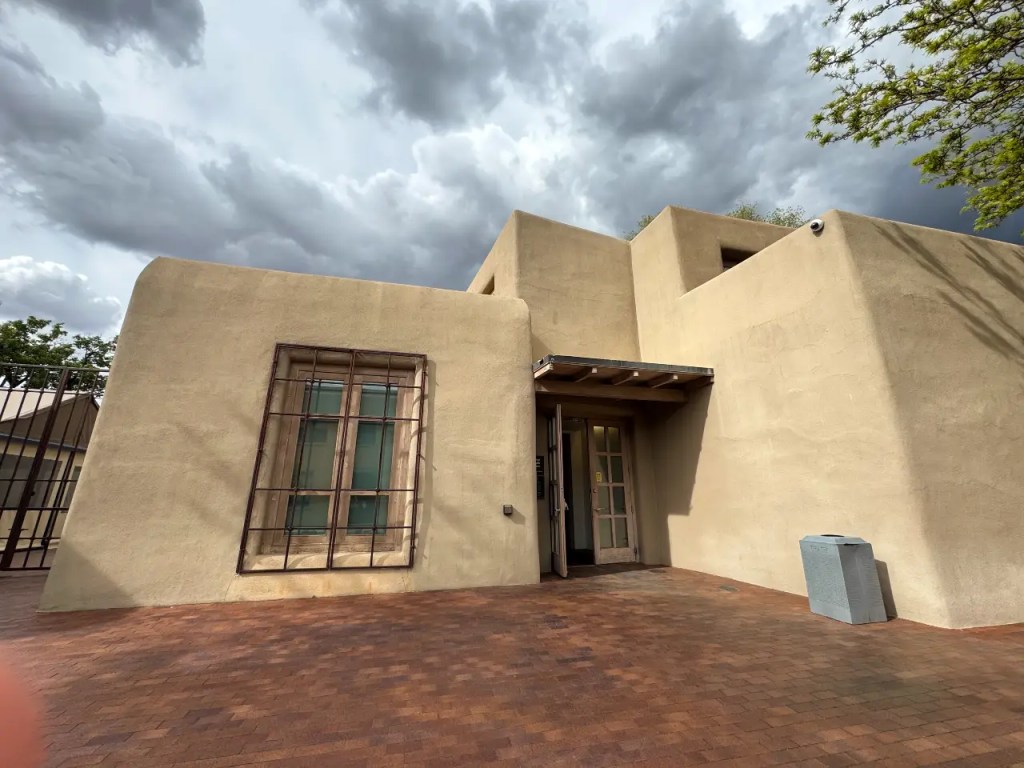 Exterior view of a southwestern style adobe building with a partially open door, surrounded by dark, cloudy skies.