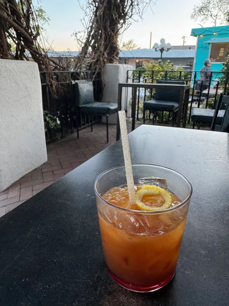 A cocktail served on a table outdoors, featuring a glass with a drink that has a slice of lemon on the rim and ice cubes, surrounded by a patio setting with greenery in the background.