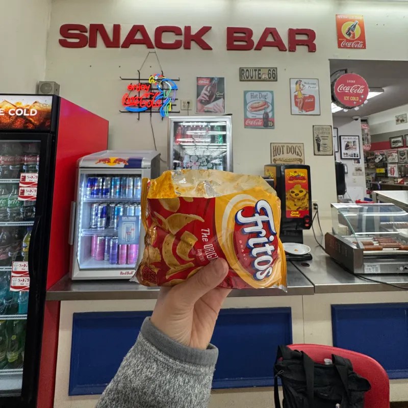 A person holding a bag of Fritos at a vintage snack bar with retro decor and a visible fridge displaying beverages.
