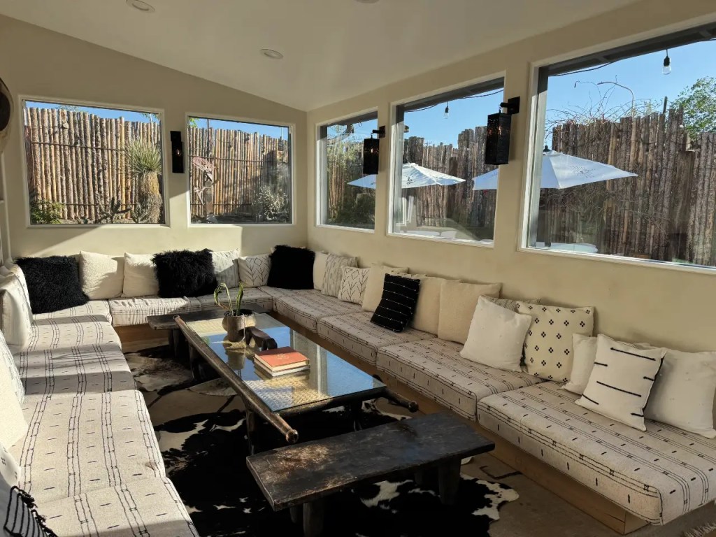 A cozy indoor seating area with a large, cushioned bench, decorative pillows, and a wooden coffee table, surrounded by windows that offer natural light and views of a fenced outdoor area.