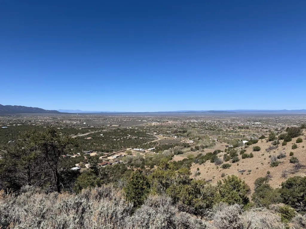 A panoramic view of a vast landscape in Northern New Mexico from the Devisadero Loop Trail, showcasing a mix of greenery and arid terrain under a clear blue sky.