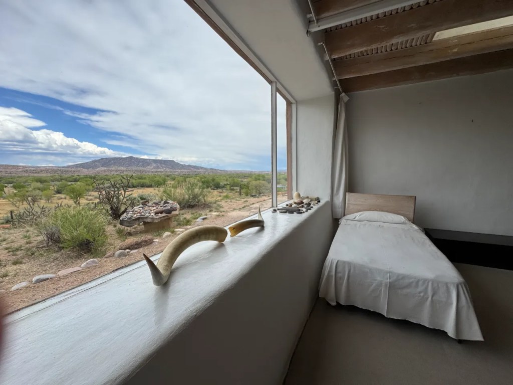 The minimalist room in Georgia O' Keefe's Abiquiu home, with a bed and decorative natural elements, showcasing a large window that opens to a scenic view of desert landscape and distant mountains.