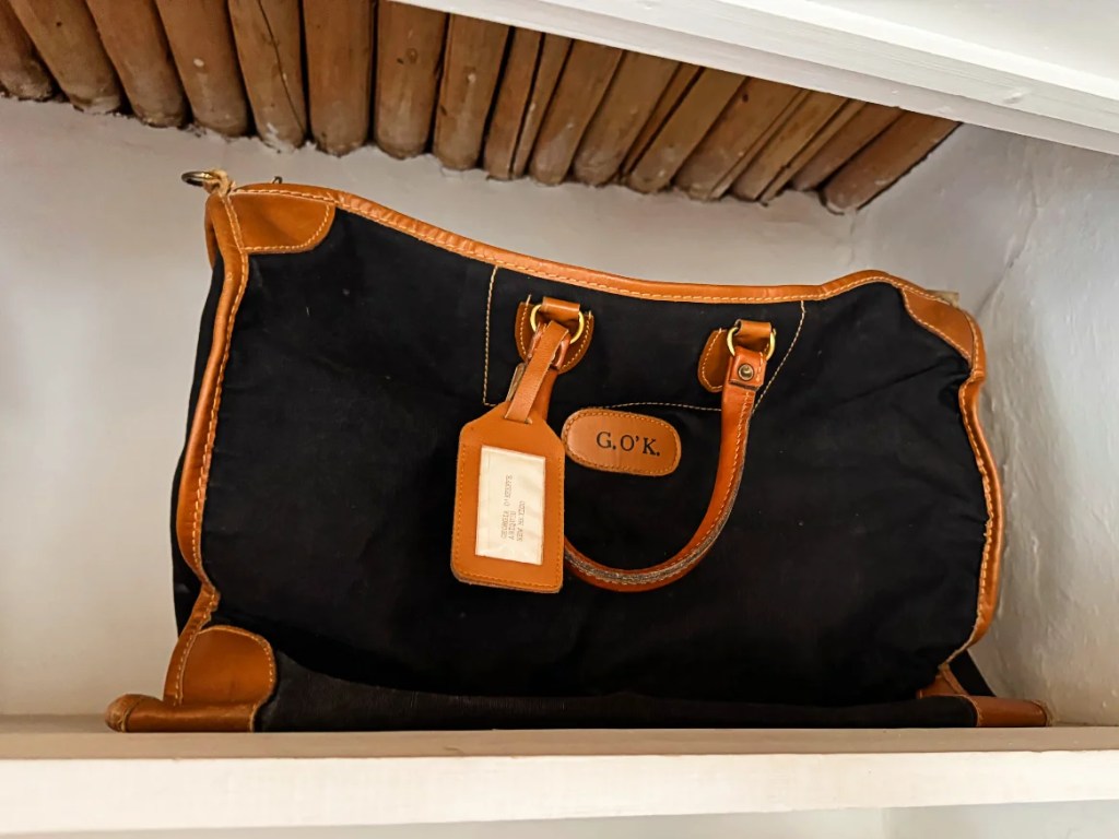 A vintage black canvas bag with leather accents, featuring monogrammed initials 'G.O.K.' and a luggage tag, displayed on a wooden shelf.