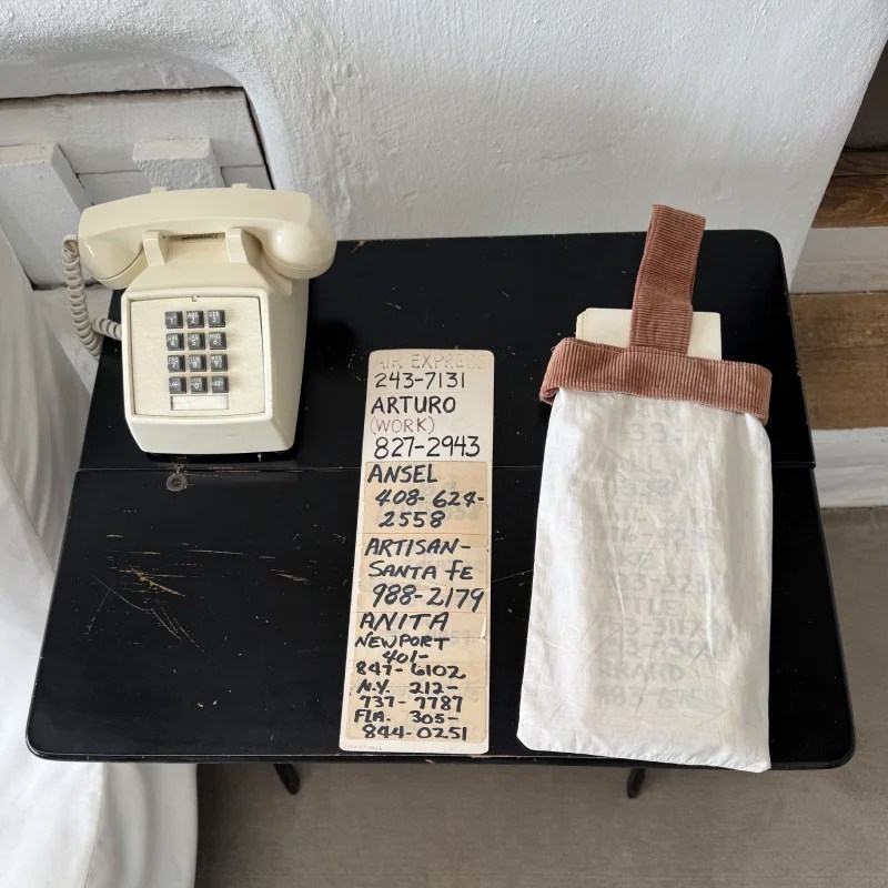 A vintage rotary phone sits on a black table alongside a notepad with contact numbers and a white fabric pouch with a brown strap.