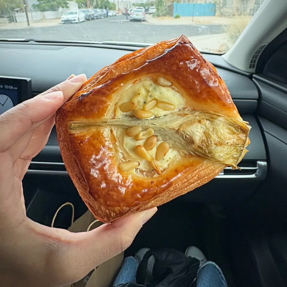 A hand holding a mango and jasmine rice danish cup with pine nuts, viewed inside a car with a street visible in the background.