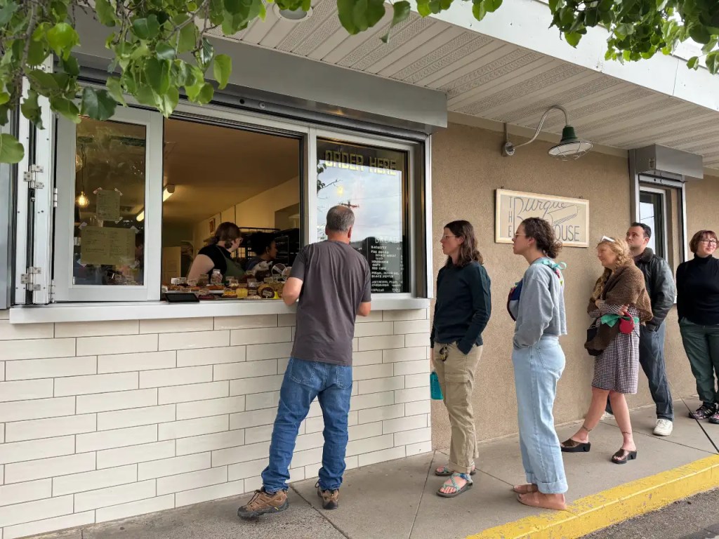 A line of customers waiting outside to order from a window at a bakery with a menu displayed outside.