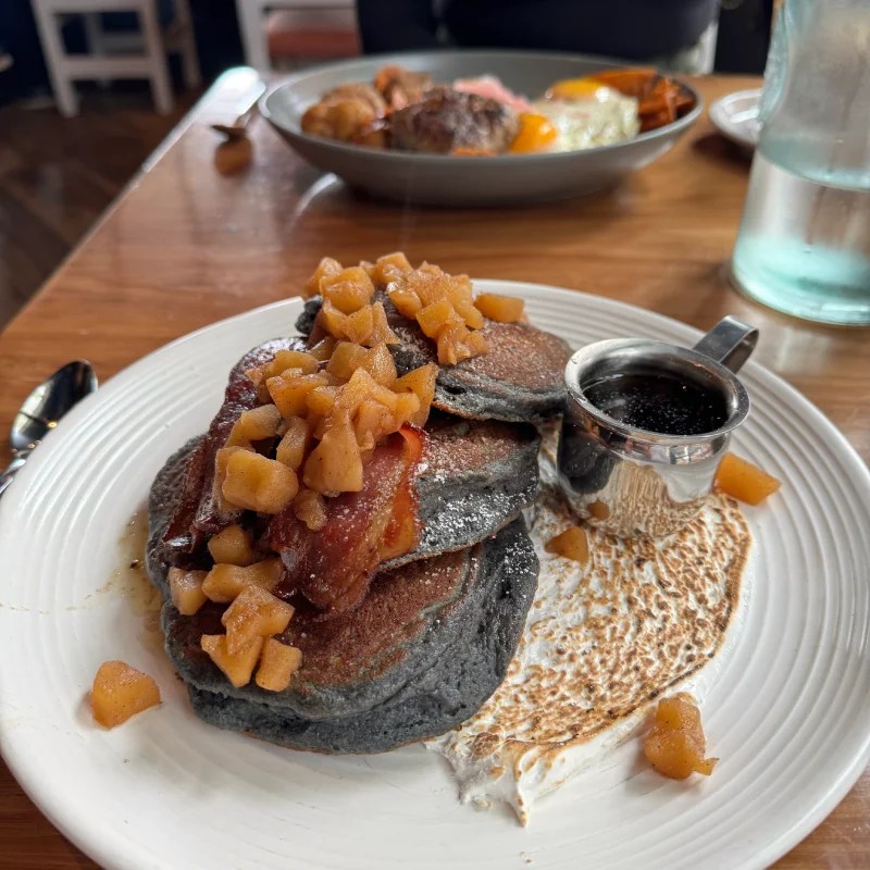 A plate of blue corn pancakes topped with sautéed apples and served with a small cup of syrup, accompanied by visible side dishes in the background.