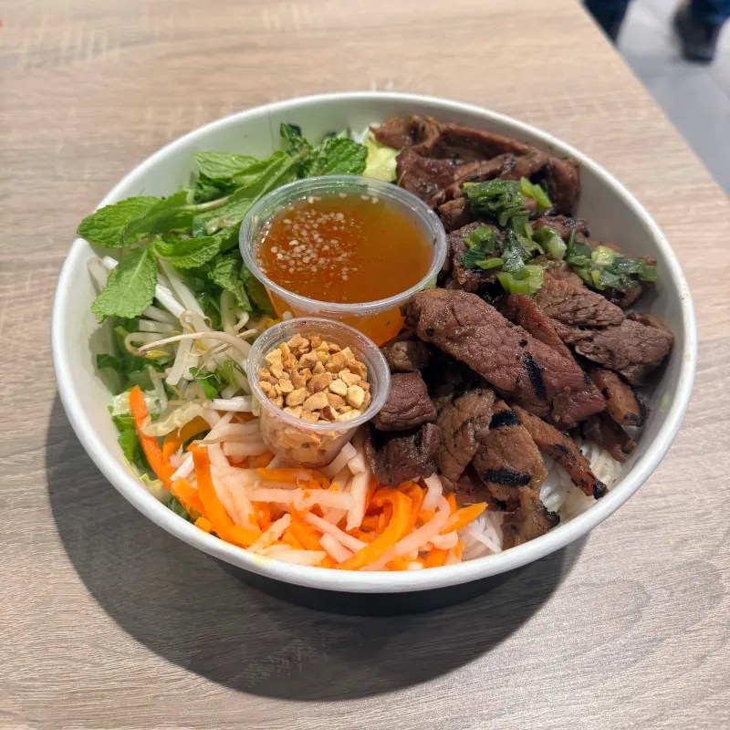 A bowl filled with grilled pork, vermicelli noodles, fresh vegetables, herbs, and a side of dipping sauce and crushed peanuts.