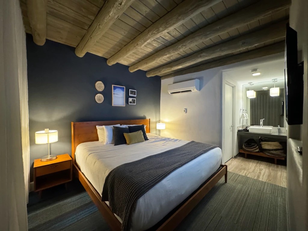 A cozy hotel room featuring a queen-sized bed, wooden furniture, and rustic ceiling beams, paired with modern amenities and a stylish bathroom in the background.