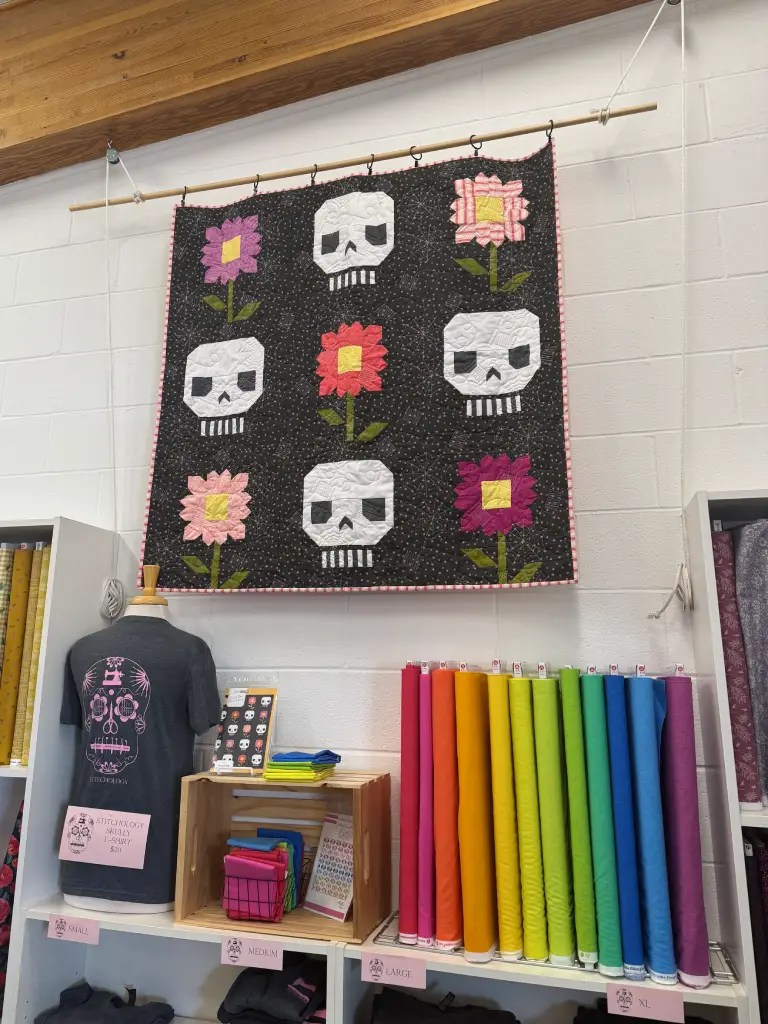 A colorful quilt featuring skulls and flowers hanging on a wall, with various fabric rolls displayed below and a skull-themed t-shirt on a mannequin.