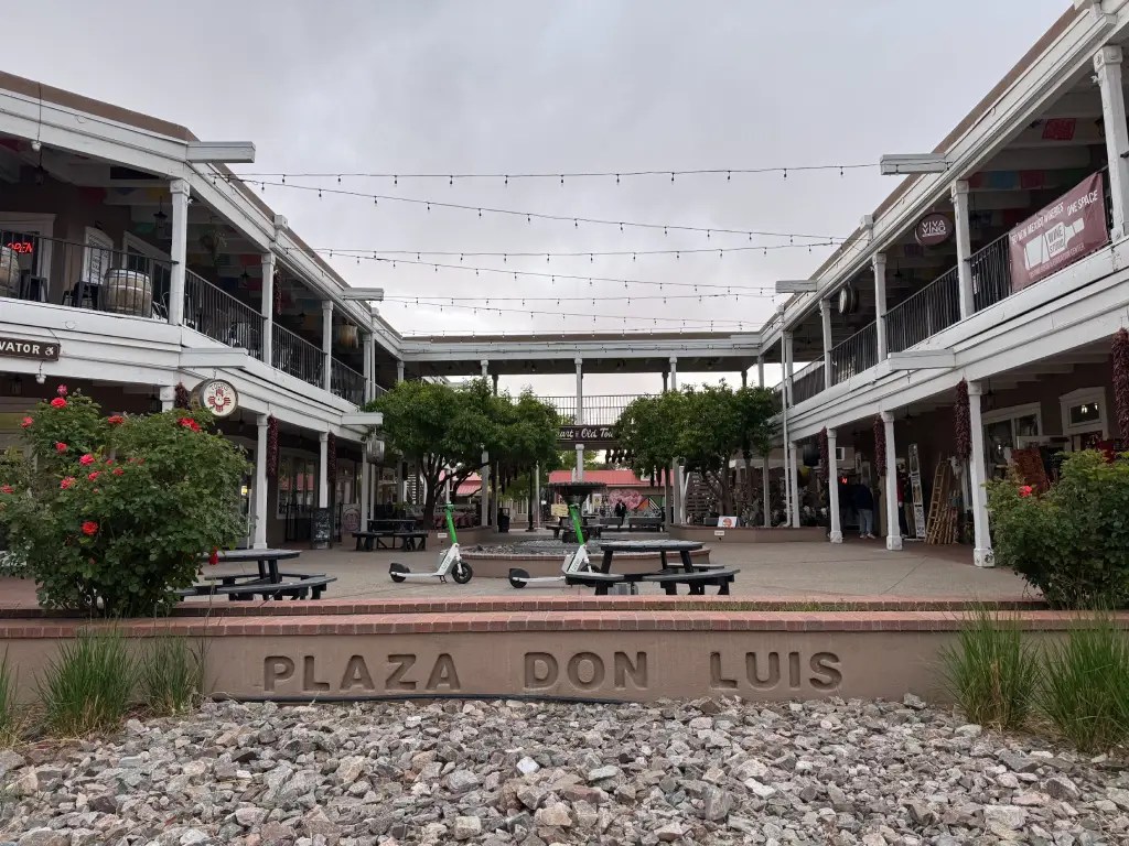 A view of Plaza Don Luis, featuring open-air shopping with string lights and small green trees. The plaza has benches and a sign embedded in the ground that reads 'PLAZA DON LUIS.'