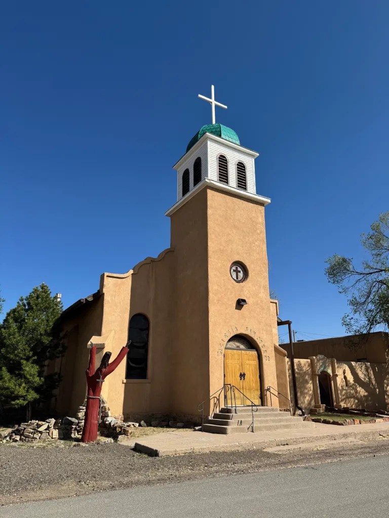 A rustic adobe church with a green domed roof and a tall cross, surrounded by trees against a clear blue sky.