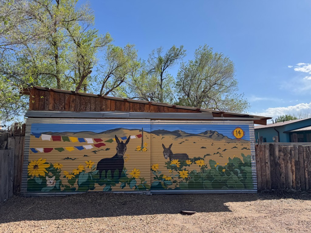 A colorful mural on a metal building featuring two donkeys amidst sunflowers and mountains, with multicolor flags fluttering in the background.