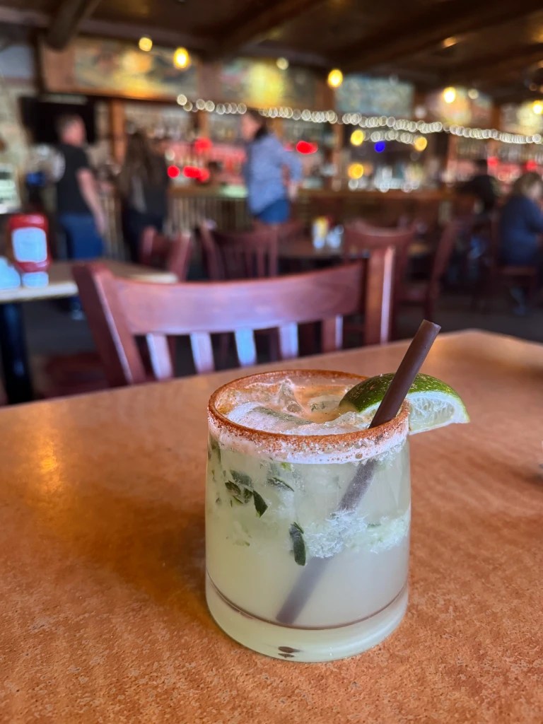 A refreshing margarita with a lime garnish and a red chile salted rim, placed on a wooden table in a lively restaurant setting.