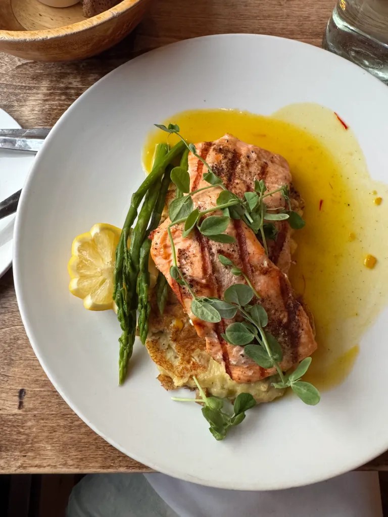 A plated dish featuring grilled salmon fillet, asparagus, and a slice of lemon, drizzled with saffron citrus sauce, served over a bed of potato cakes.