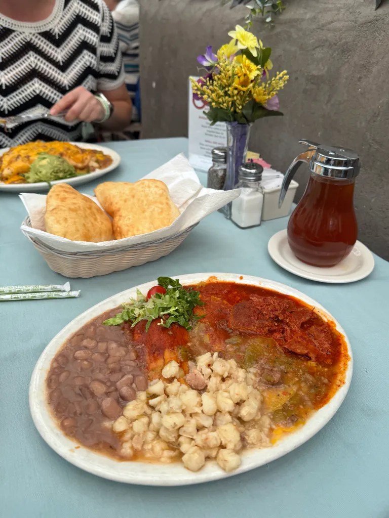A plate of traditional New Mexican food featuring enchiladas topped with red and green sauces, accompanied by refried beans, posole, and a garnish of lettuce and tomato. In the background, a person is seated at a table, and there is a small bouquet of flowers.