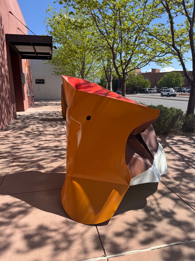 A colorful abstract sculpture displayed outdoors, featuring vibrant orange, red, and brown sections, surrounded by trees and a sidewalk.