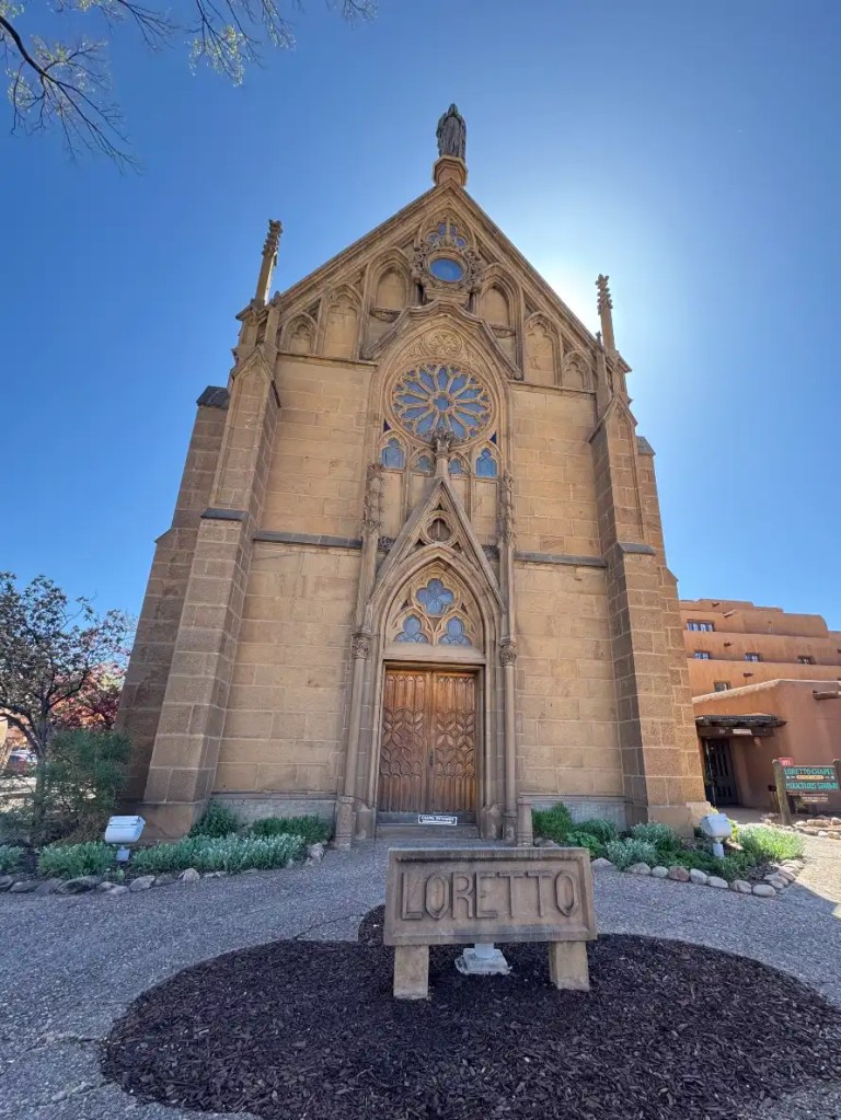 Front view of Loretto Chapel in Santa Fe, New Mexico, showcasing its Gothic architectural style, rose window, and a statue atop the building under a clear blue sky.
