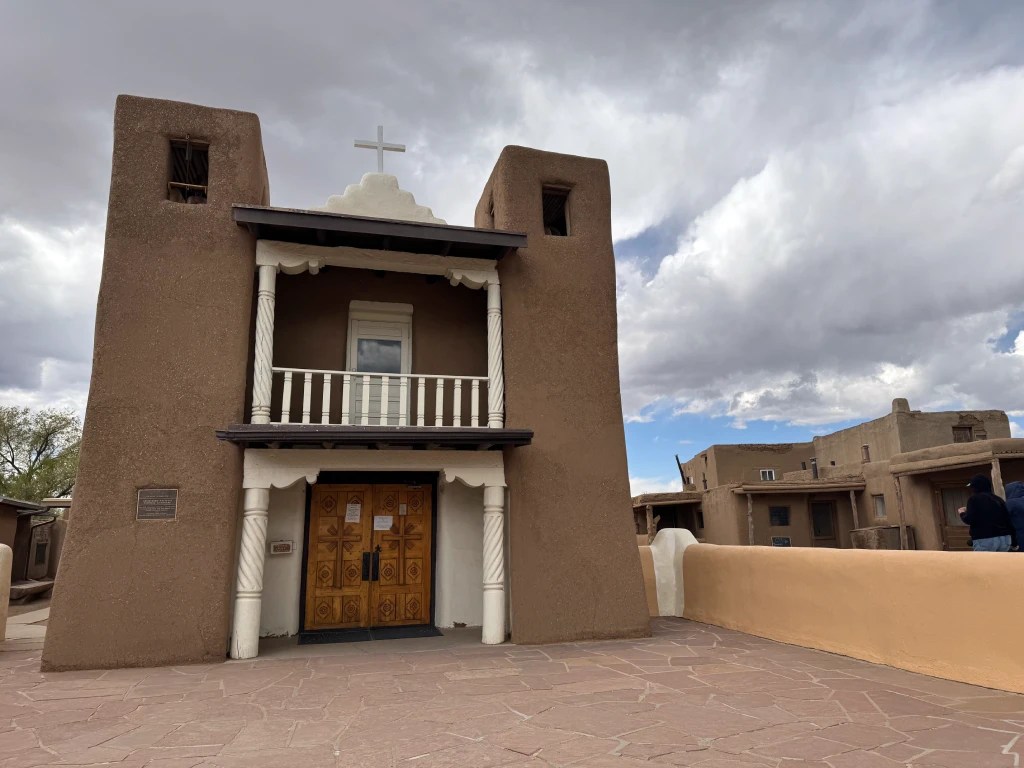 San Geronimo church in Taos Pueblo, featuring a cross on top, brown walls, a decorative wooden door, and a balcony, with a cloudy sky in the background.