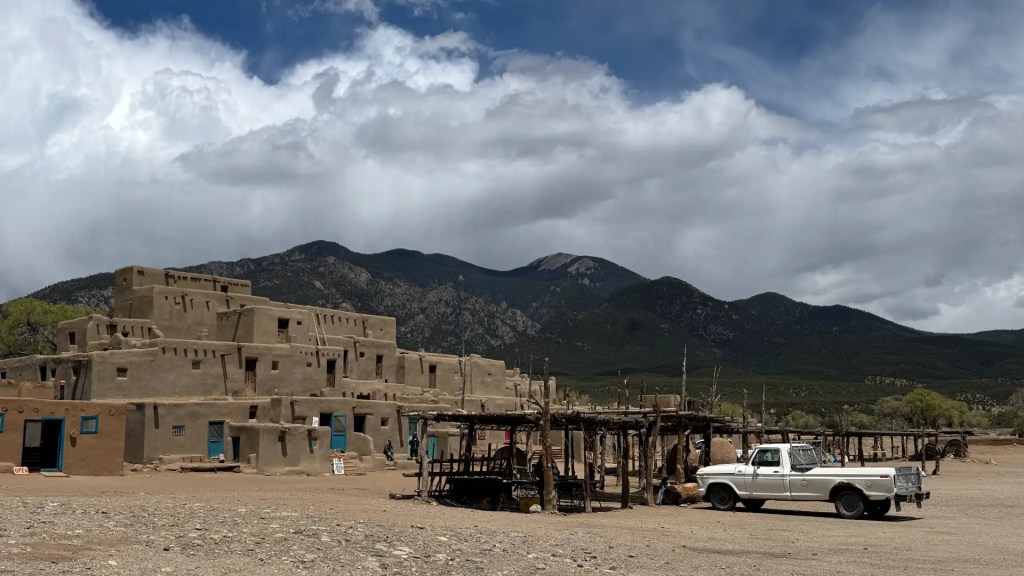 Traditional adobe buildings of Taos Pueblo against a backdrop of mountains and cloudy skies, with a parked white truck in the foreground.
